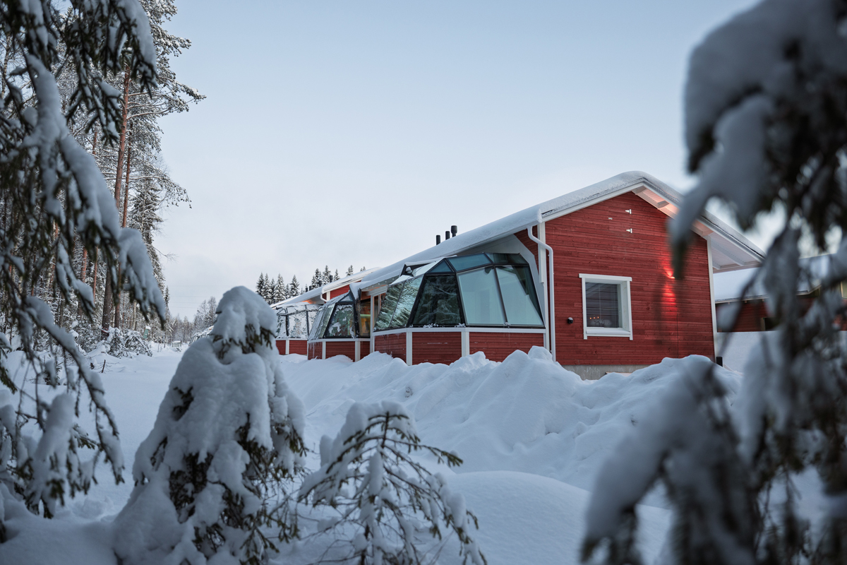 Red-painted glass igloo cottage with a glass roof section set among snow-covered trees at dusk, Santa Claus Holiday Village, Rovaniemi