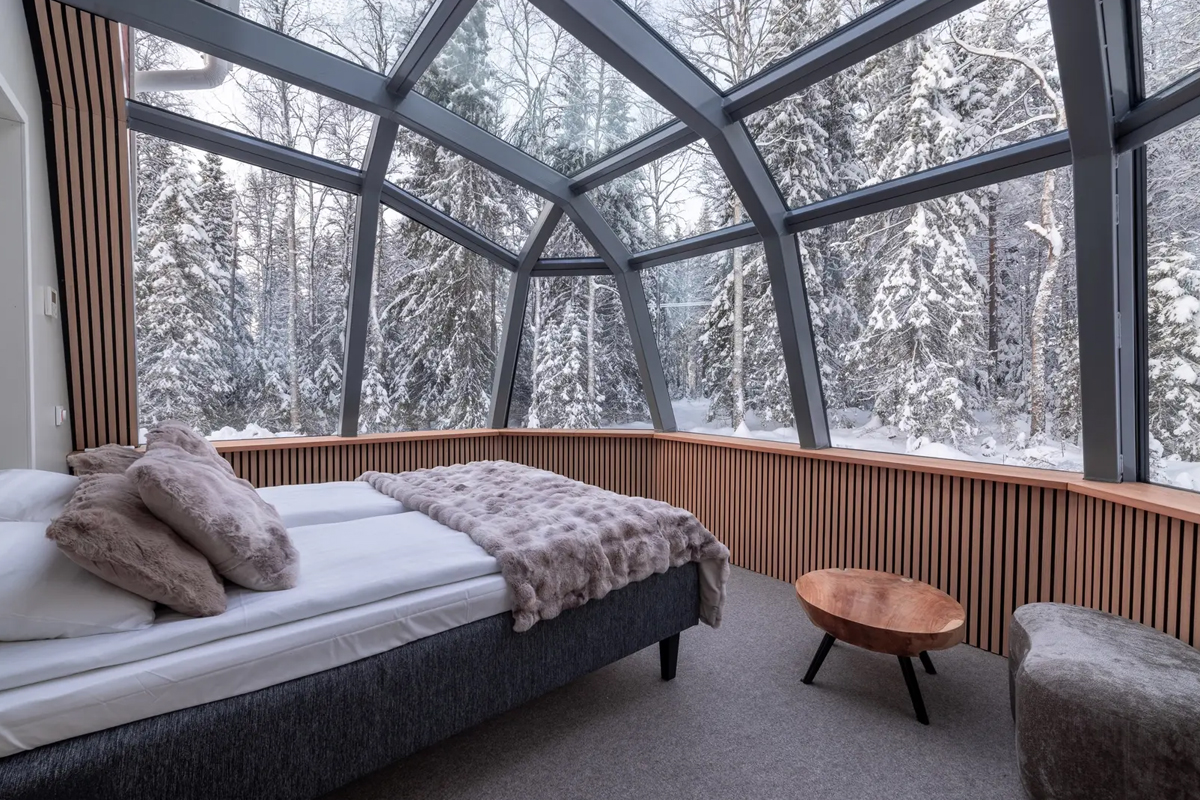 Glass igloo sleeping area with a bed under a full glass ceiling overlooking snow-covered trees, Santa Claus Holiday Village, Rovaniemi