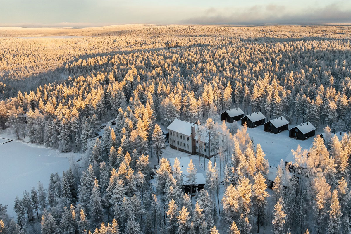 Aerial winter view of Skýra Retreat and its log cabin accommodation, set within a snow-covered forest beside Lake Norvajärvi.