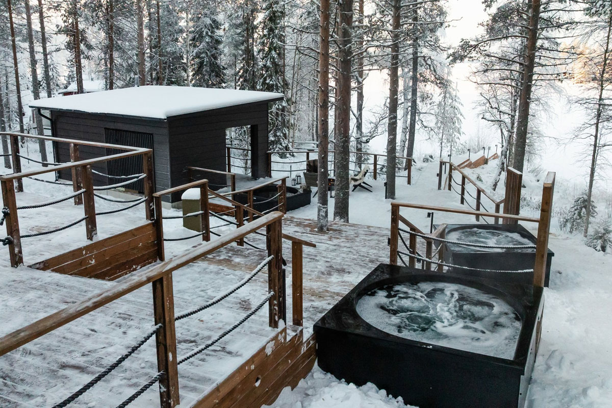 Bains à remous extérieurs et bâtiment du sauna au spa nordique du Skýra Retreat, entourés d'arbres enneigés près de Rovaniemi.