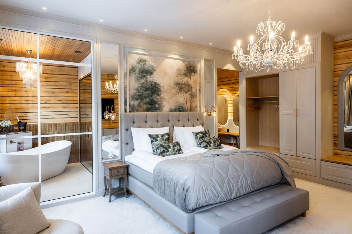 Chambre d'une Grande Suite du Skýra Retreat avec lit artisanal, murs en rondins, lustre et salle de bain privée avec baignoire et sauna.