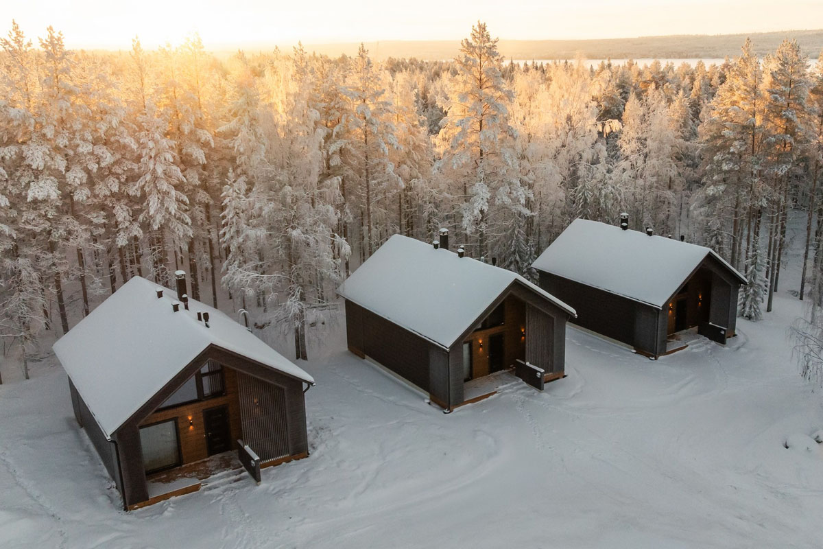 Trois suites en rondins du Skýra Retreat au milieu des arbres enneigés, avec le lac Norvajärvi visible en arrière-plan au coucher du soleil.