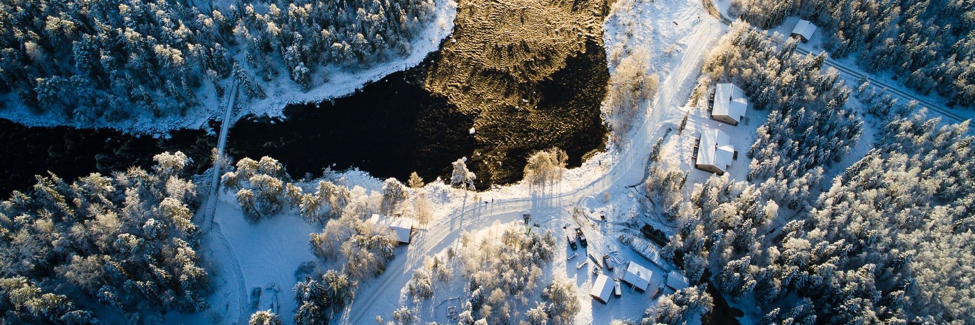 Vue aérienne du Vaattunki Wilderness Resort au bord de la rivière en hiver, avec les hébergements et la forêt visibles dans le paysage enneigé près de Rovaniemi.