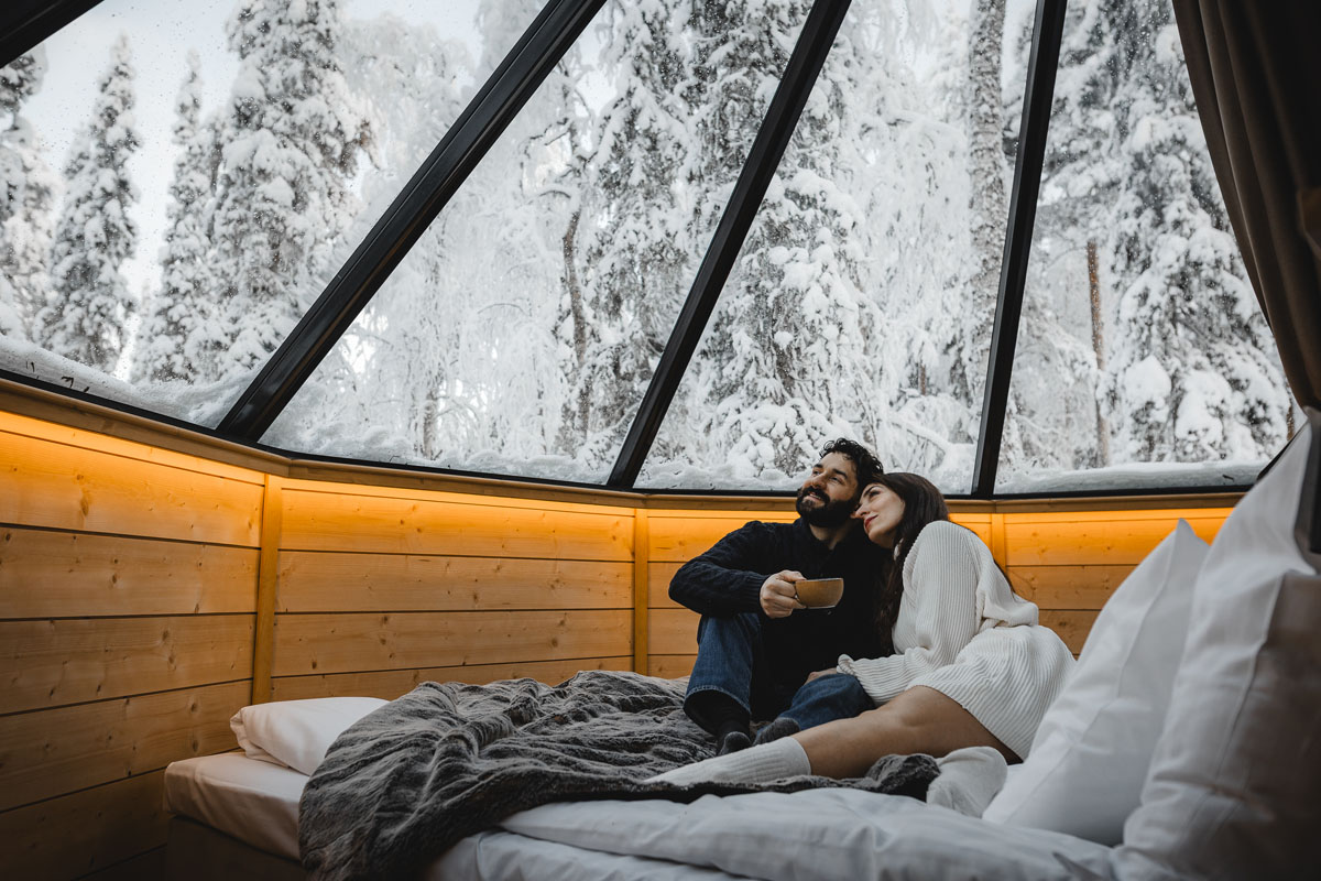 Couple se détendant à l'intérieur d'un Aurora Cabin Igloo au Vaattunki Wilderness Resort, contemplant la forêt enneigée à travers le toit en verre.