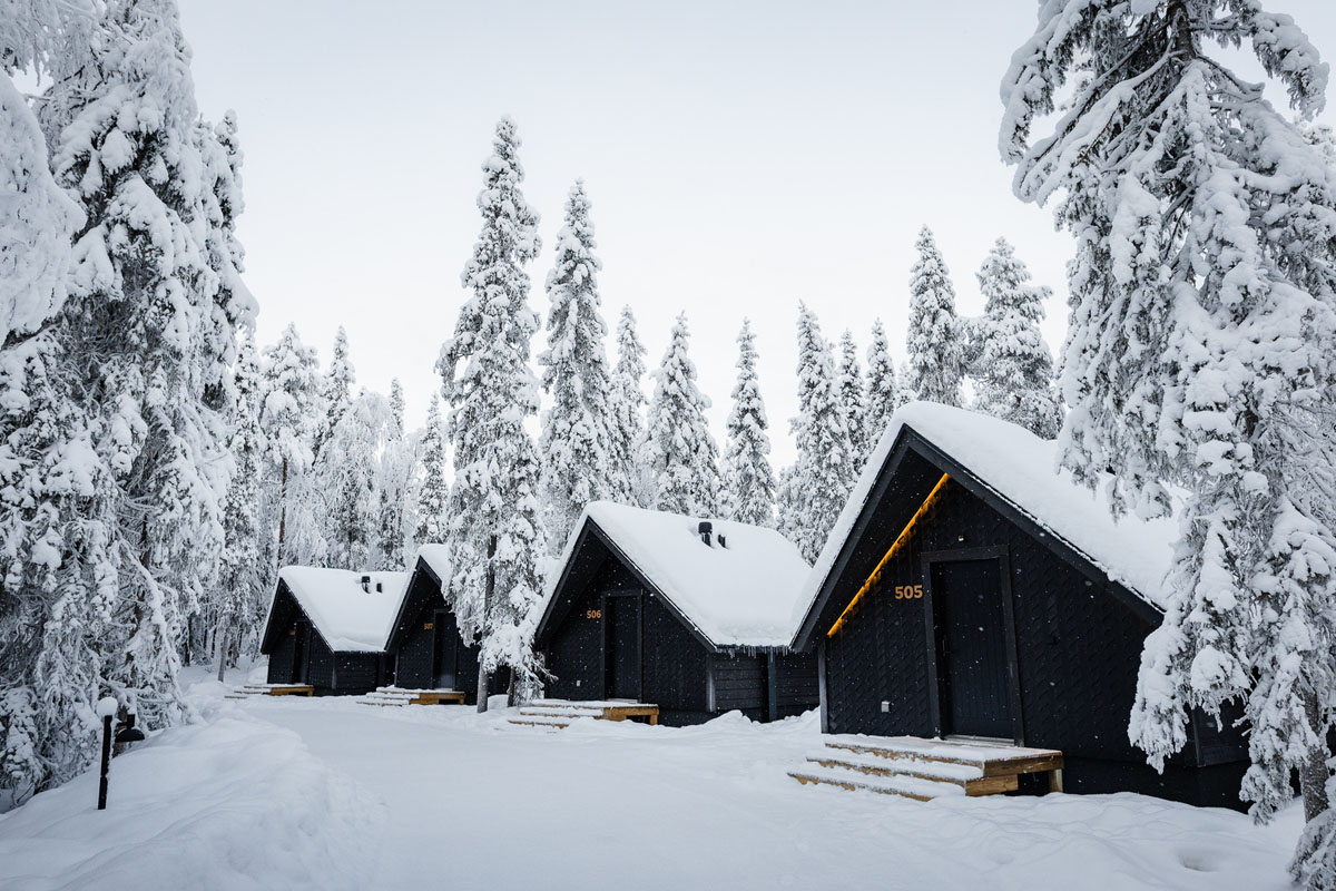 Rangée de chalets Panorama Hut au Vaattunki Wilderness Resort sous une forte chute de neige, entre de grands arbres enneigés près de Rovaniemi.