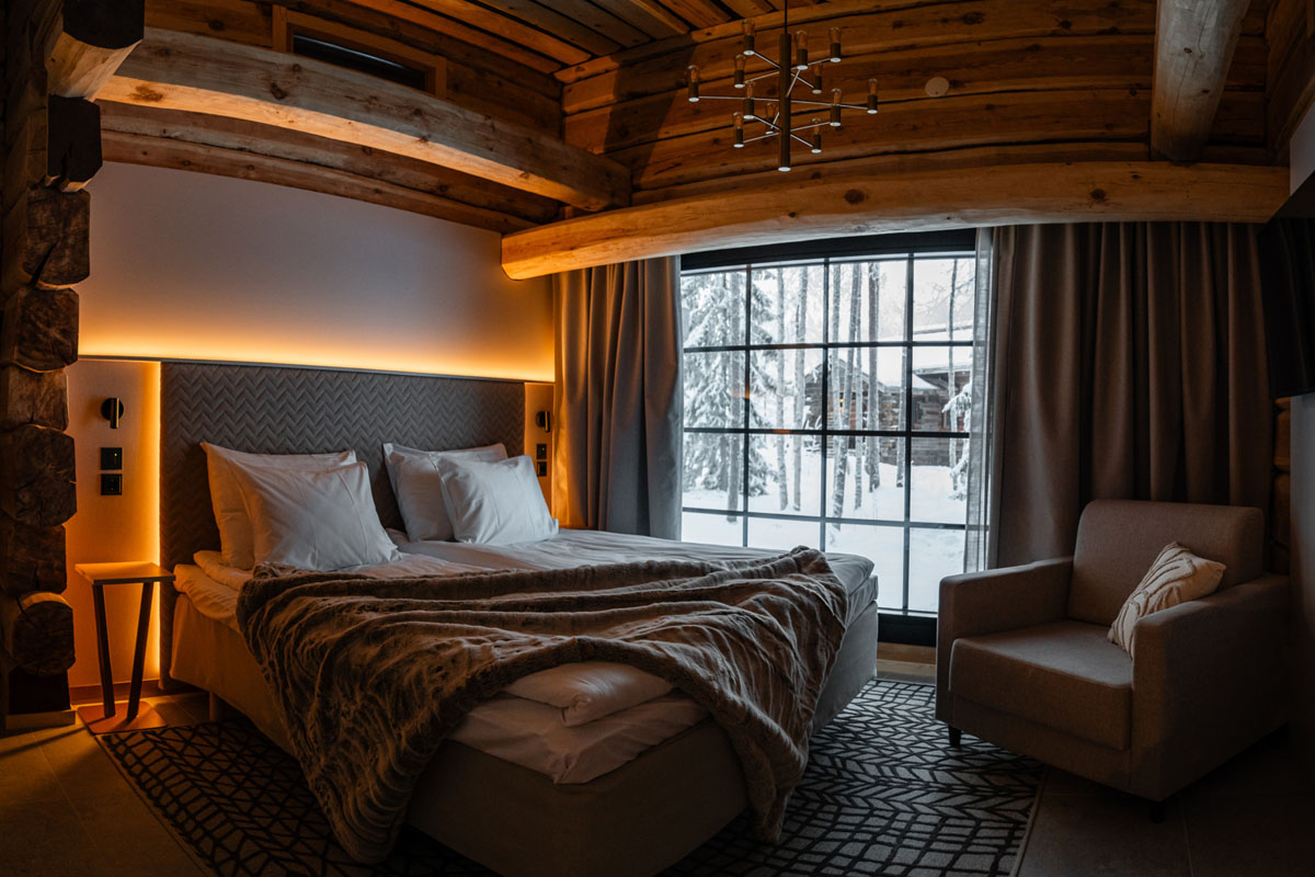 Chambre d'une Arctic Deluxe au Vaattunki Wilderness Resort, avec poutres en rondins au plafond, éclairage chaleureux et fenêtre donnant sur la forêt.