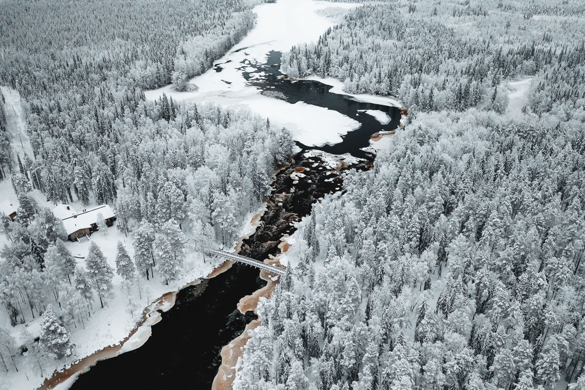 Vue aérienne hivernale de la rivière et de la forêt environnante au Vaattunki Wilderness Resort près de Rovaniemi, avec une passerelle visible en contrebas.