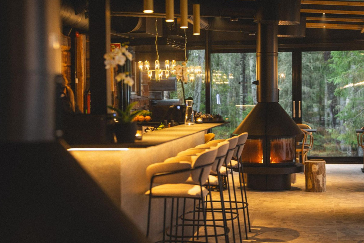 Bar et salon du restaurant Niva au Vaattunki Wilderness Resort, avec feu ouvert et vue sur la forêt à travers de grandes baies vitrées.