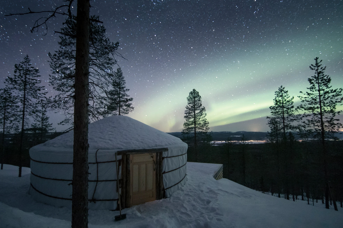 A snow covered yurt at Yurt District glowing under the northern lights and a starry sky.