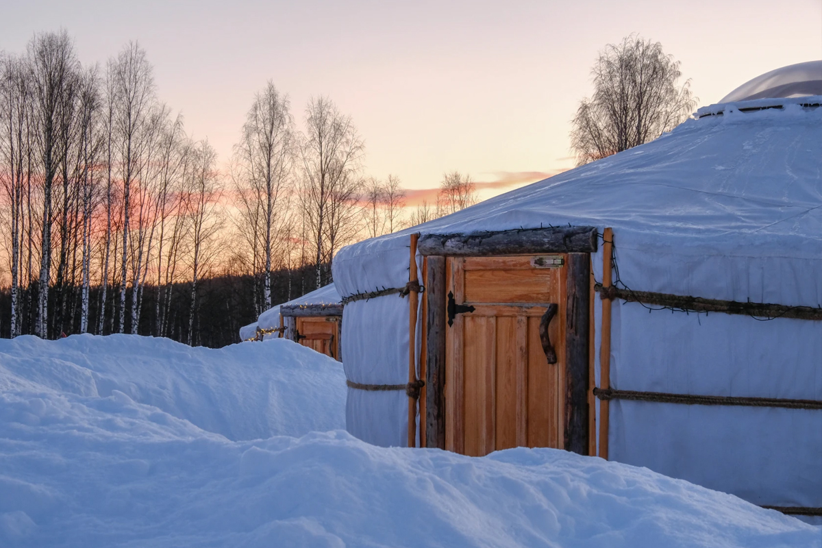 Snow covered yurts at Yurt District with wooden doors, lit by a pink winter sunset.
