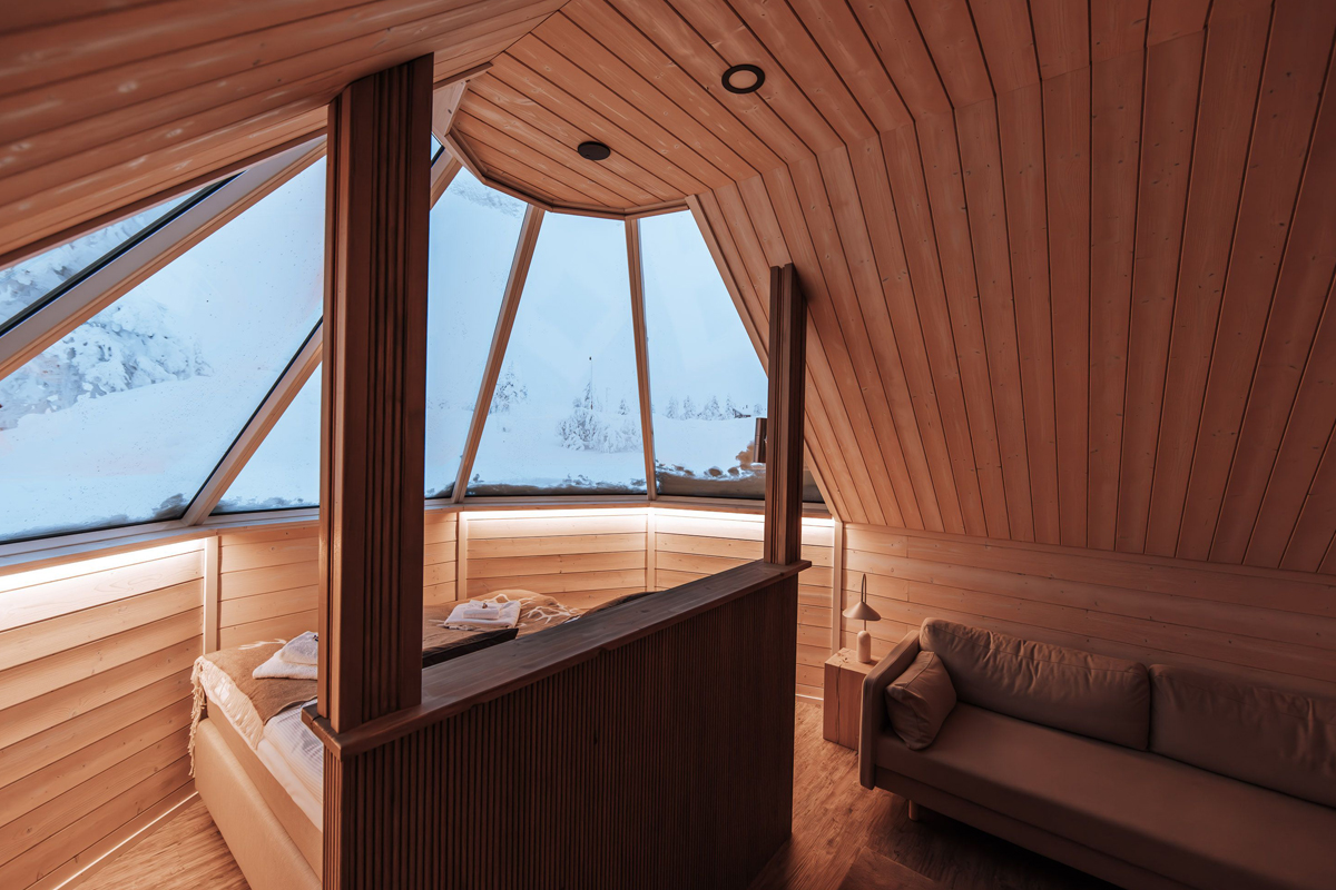 Curved wood-paneled aurora cabin bedroom with a panoramic roof window overlooking a snowy slope