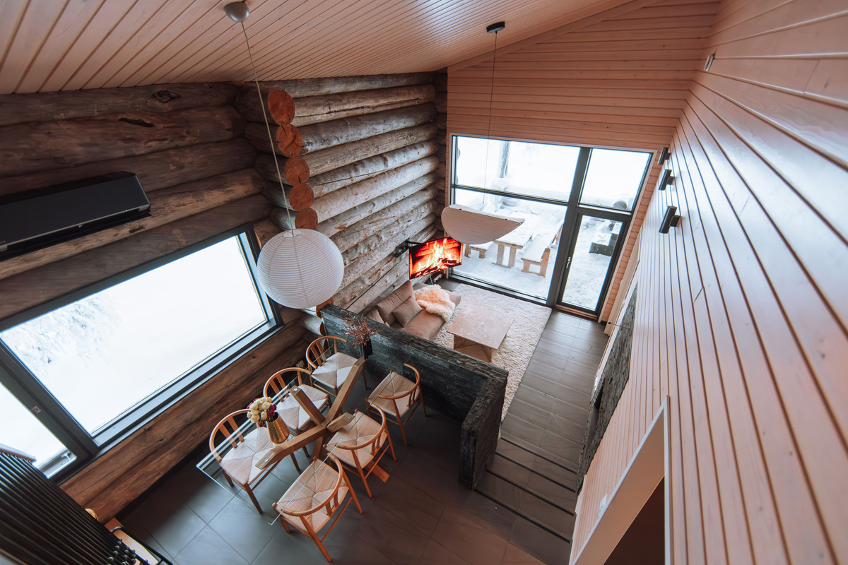 Overhead view of a cabin interior with a lit fireplace, dining area, and floor-to-ceiling windows facing a snowy garden