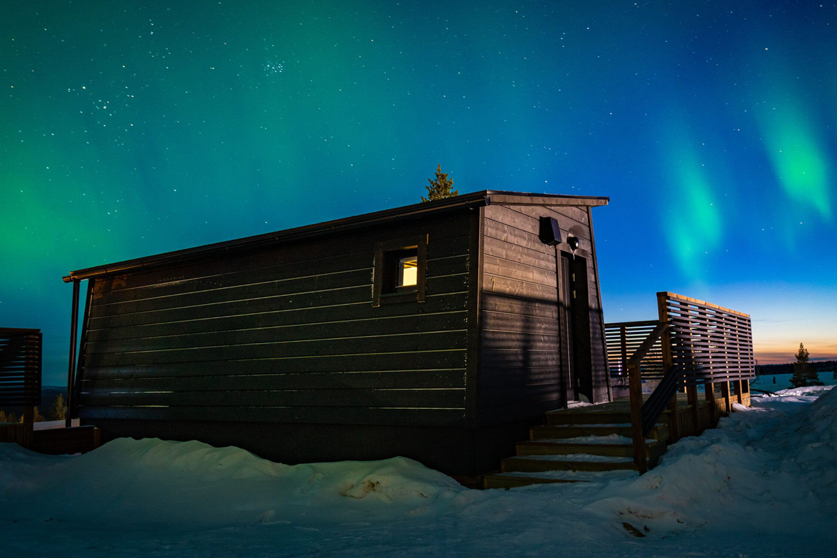 Aurora Collection cabin at twilight with green and teal northern lights across a star-filled sky, Saariselkä