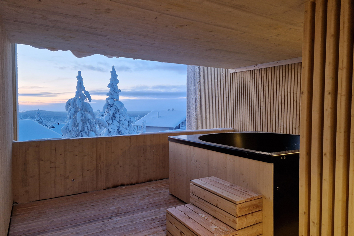 Private terrace with a black outdoor bathtub on a wooden deck, overlooking snow-covered trees and a pale winter sky
