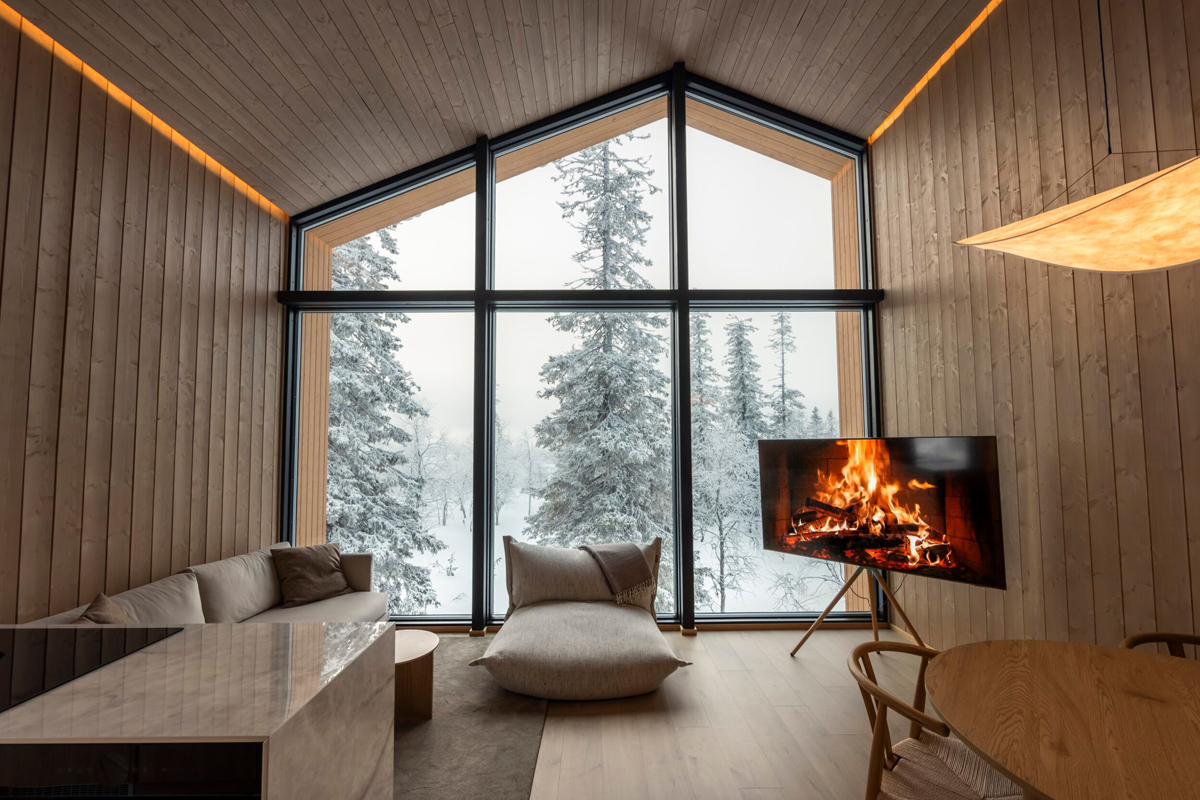 Bright cabin lounge with a gabled glass wall, floor-length windows, a wood stove, and a view of a snowy forest