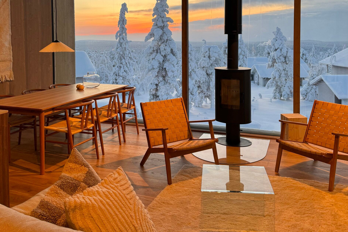 Modern cabin living room with a wood-burning stove, Scandinavian furniture, and floor-to-ceiling windows framing a sunset over snowy trees
