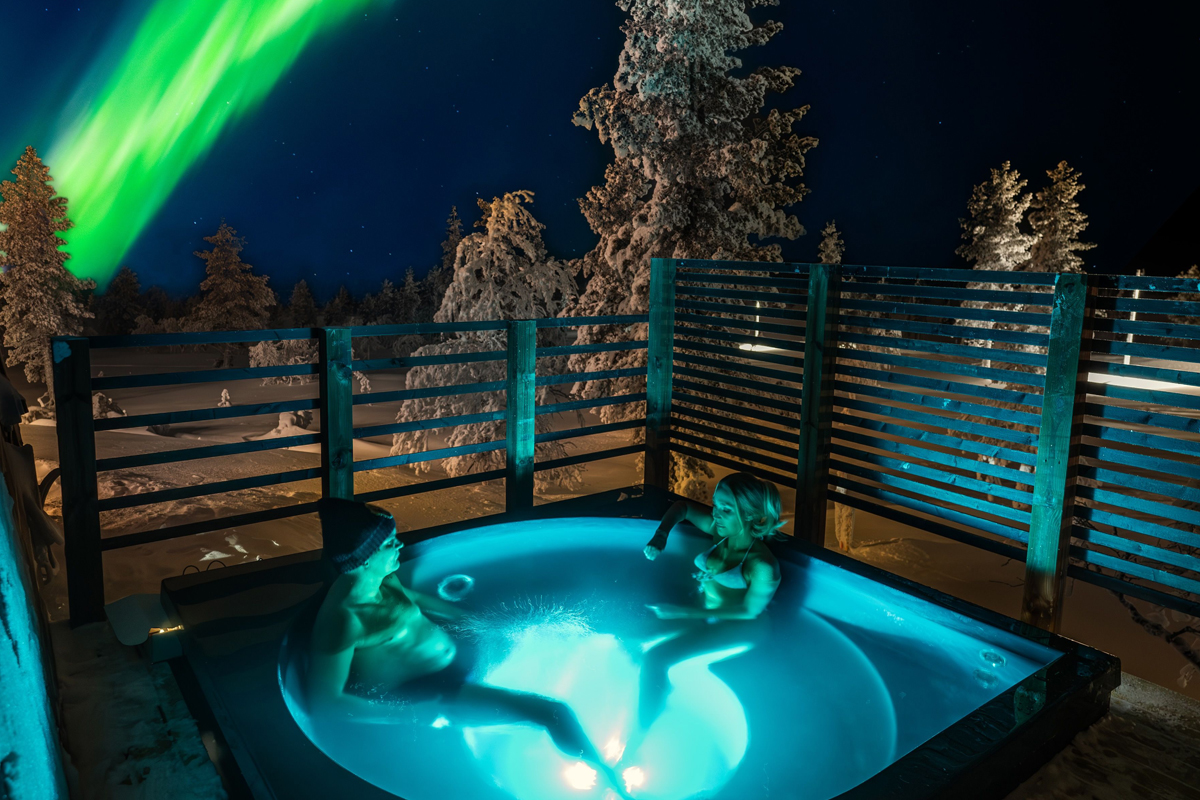 Two guests relaxing in a glowing outdoor jacuzzi at night with northern lights visible above the snowy forest