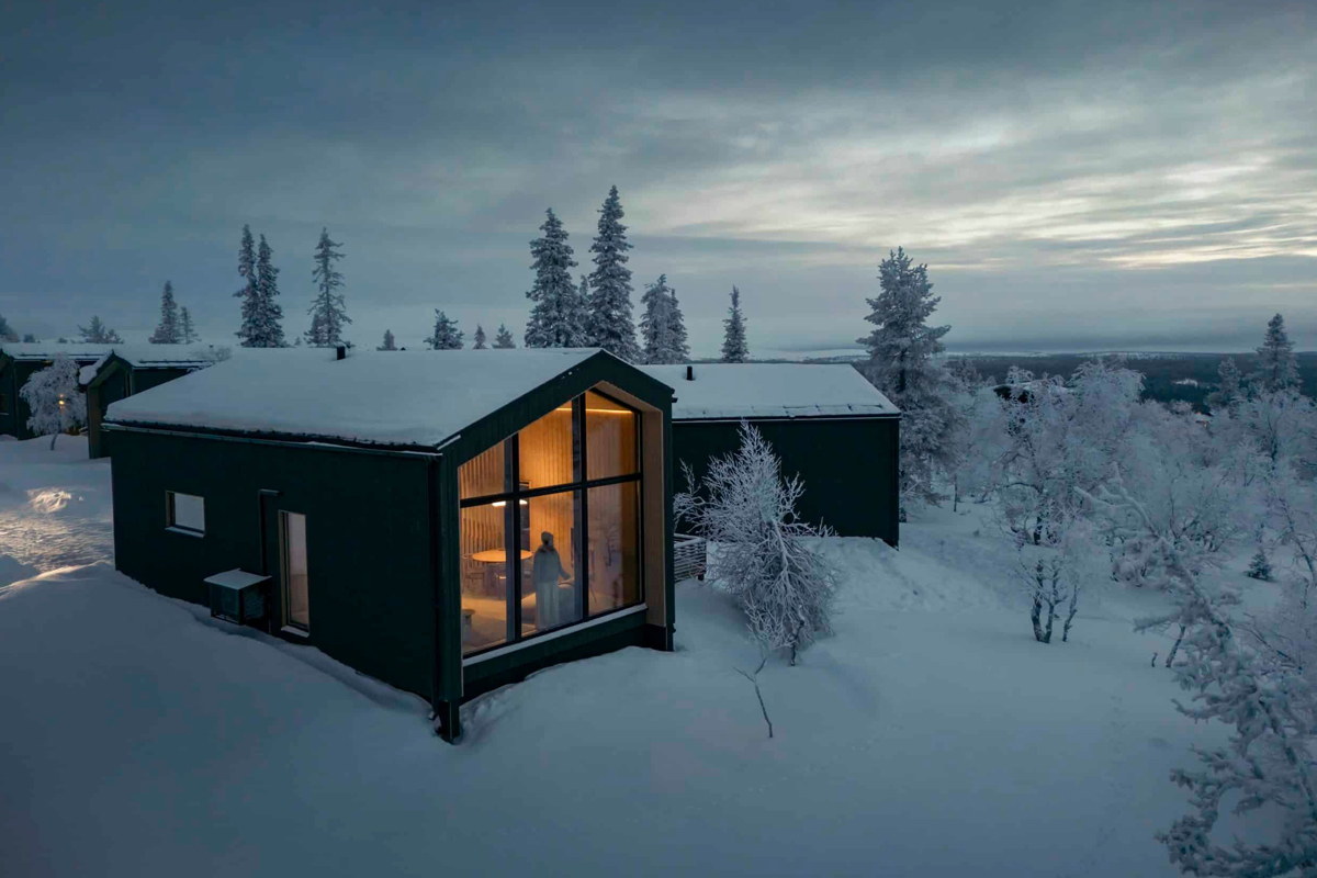 Dark green cabin glowing from within at dusk, set among frost-covered birch and pine trees in winter, Saariselkä