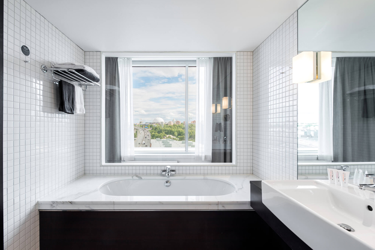 Bathroom with bathtub and city views at Clarion Hotel Stockholm in Södermalm, with white tiled walls and a large window.