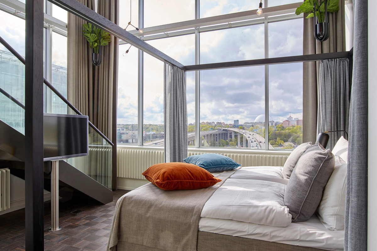 Etage Suite at Clarion Hotel Stockholm with a double-height glass facade, city views, and a loft-style bedroom with colourful cushions.