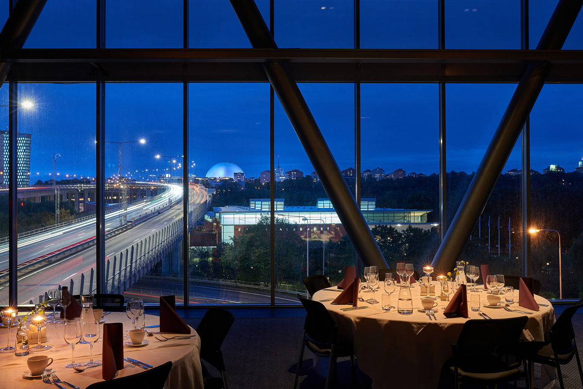 Dining area at Clarion Hotel Stockholm at night, with panoramic floor-to-ceiling windows overlooking the Stockholm cityscape and the Avicii Arena.