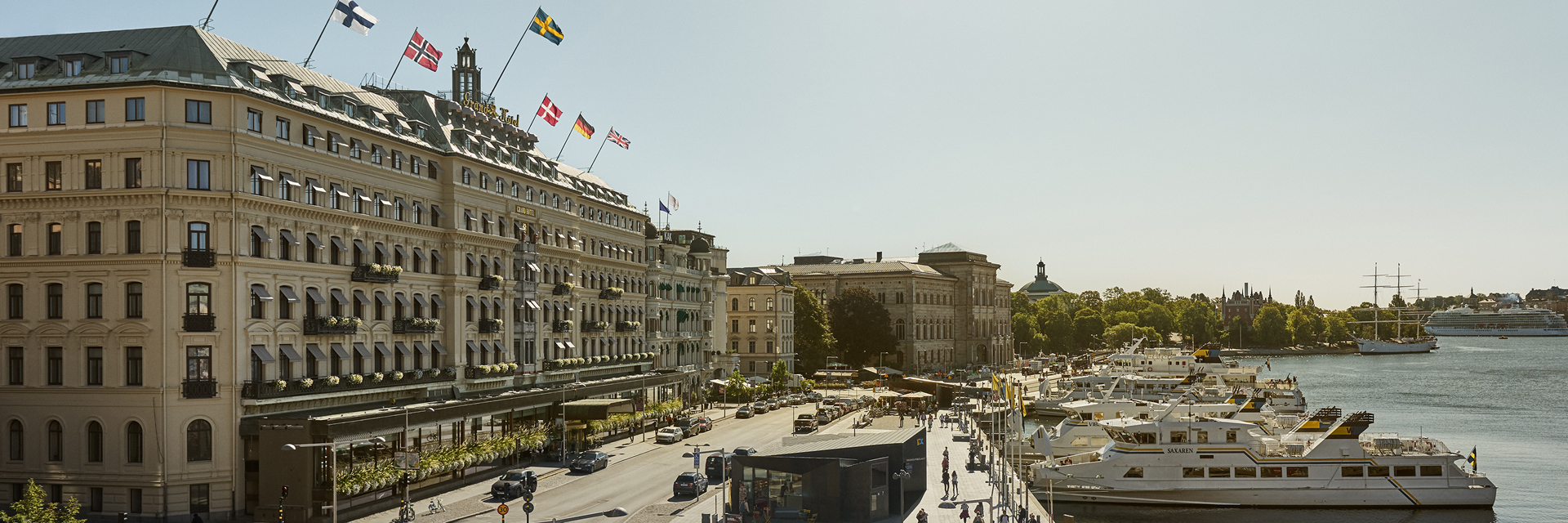 Facade of Grand Hôtel Stockholm seen from the waterfront in summer, with flags flying and boats docked along the quay.