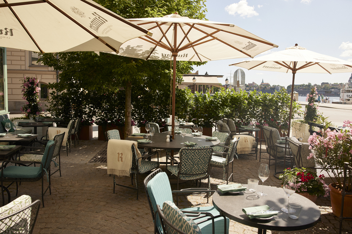 Waterfront terrace at Grand Hôtel Stockholm in summer, with Ruinart parasols, green seating, and views across the water toward Gamla Stan.