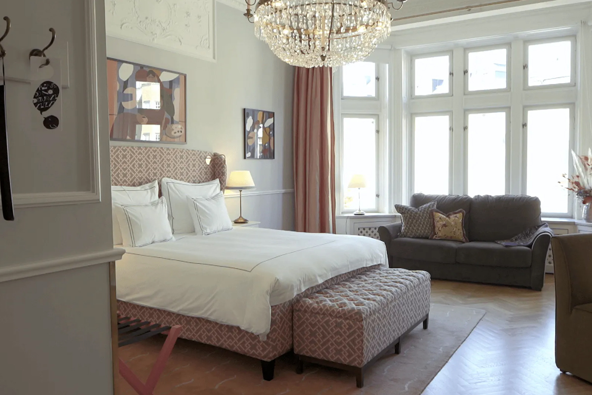 Deluxe room at The Sparrow Hotel in Stockholm, with a king-size bed, crystal chandelier, sitting area, and large windows overlooking the street.