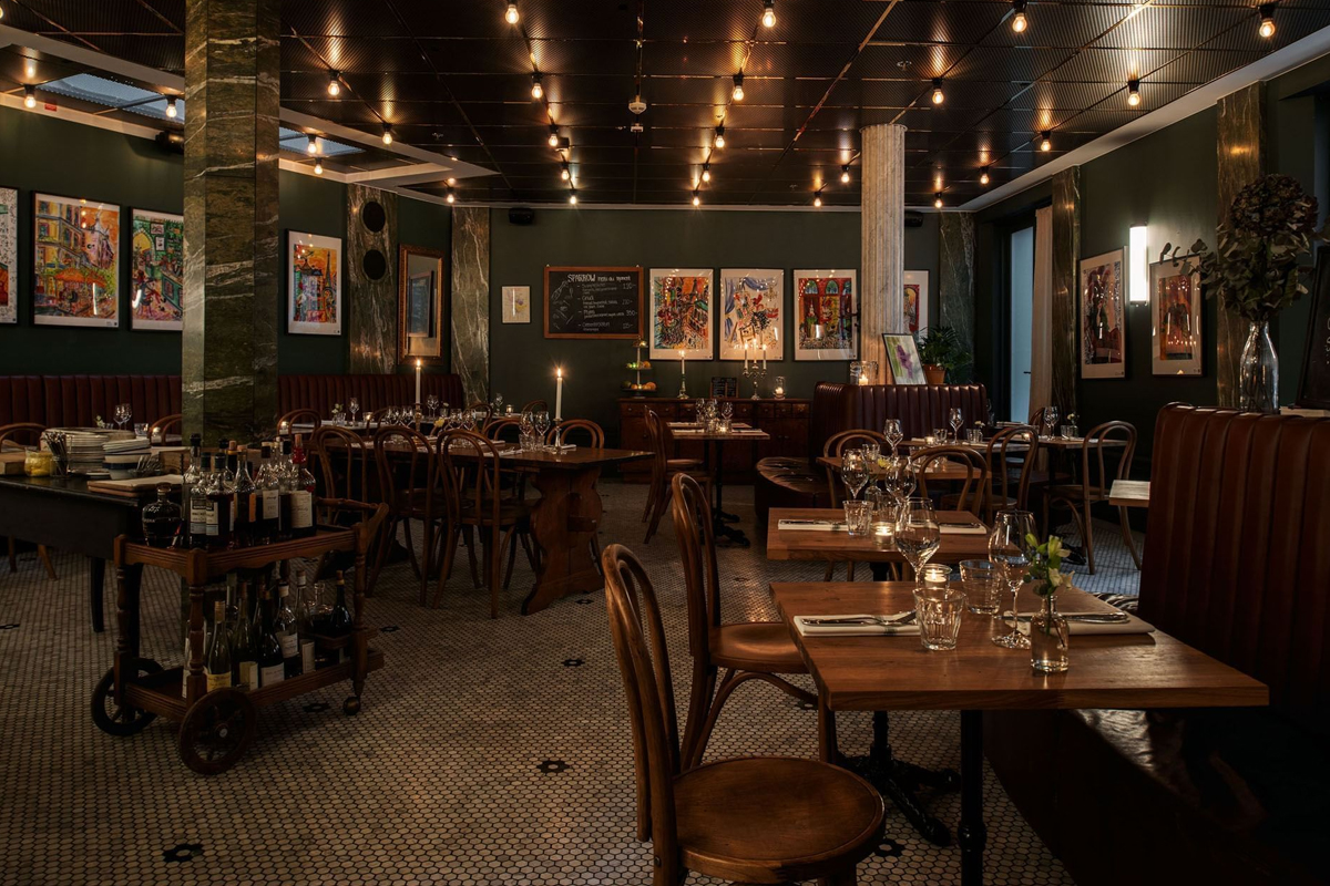 Interior of the bistro at The Sparrow Hotel in Stockholm, with wooden bistro chairs, candlelit tables, green walls, and colourful artwork.