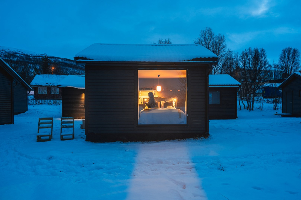 Aurora Glass Cabin at Aera in Tromso, lit up at dusk in the snow.