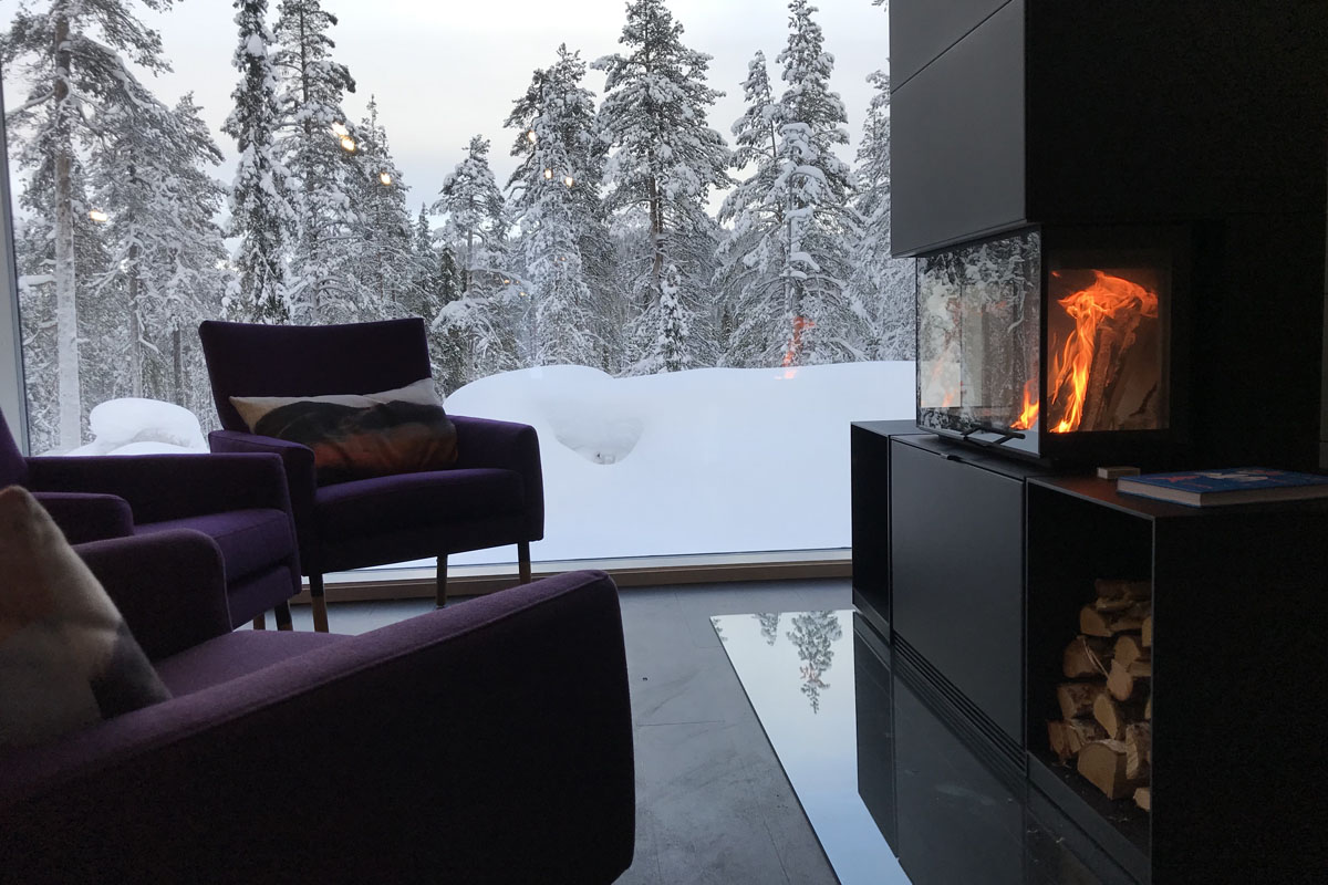 Sitting area with fireplace and panoramic forest views inside a glass cabin at Arctic Skylight Lodge near Ylläs, Finnish Lapland.