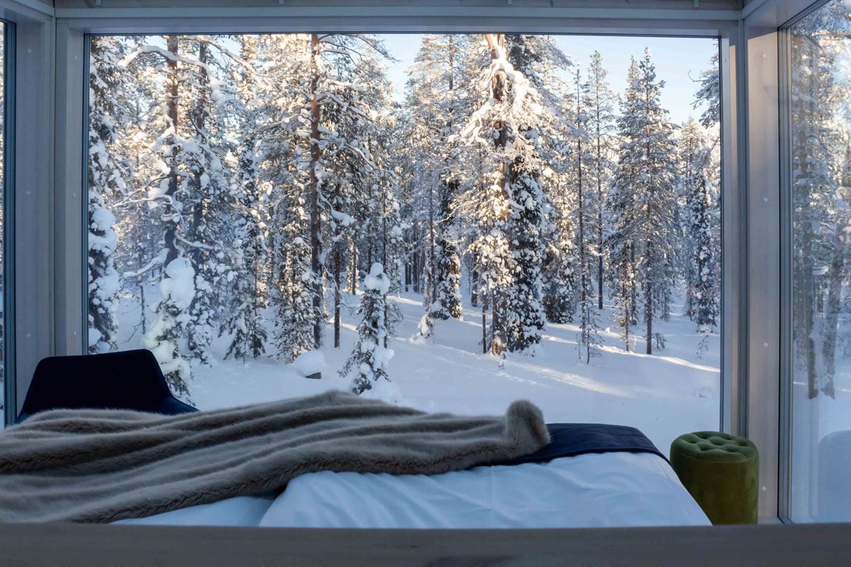 Bed with forest view through floor-to-ceiling windows in a glass cabin at Arctic Skylight Lodge near Ylläs in winter.