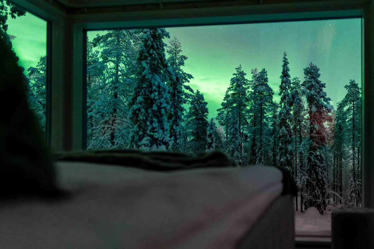 Northern lights visible through the glass wall of a cabin at Arctic Skylight Lodge near Ylläs, with snow-covered trees in the foreground.