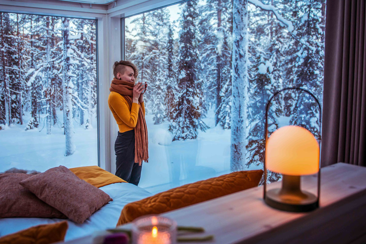 Guest looking out through the panoramic glass wall of a cabin at Arctic Skylight Lodge near Ylläs, Finnish Lapland.