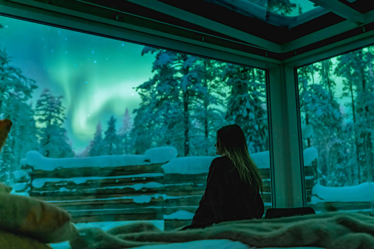 View from inside a glass cabin at Arctic Skylight Lodge near Ylläs, with the northern lights visible through the panoramic windows.