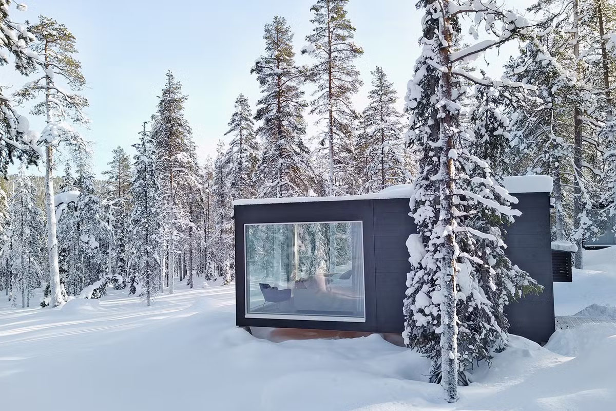 Exterior of a glass cabin at Arctic Skylight Lodge in winter, with large panoramic windows facing the snow-covered forest near Ylläs.