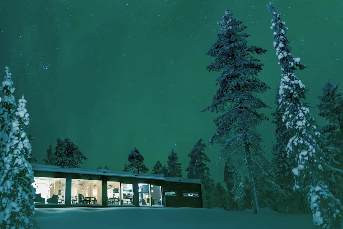 Glass-walled restaurant at Arctic Skylight Lodge lit up at night under the northern lights near Ylläs, Finnish Lapland.