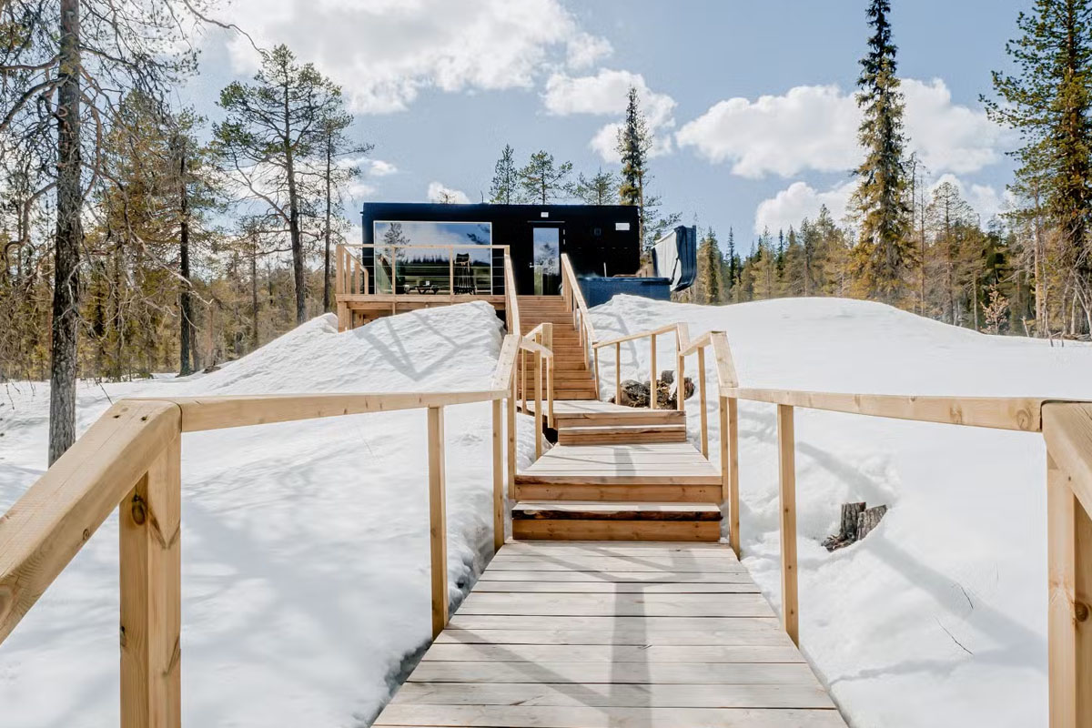 Wooden walkway leading to a glass cabin at Arctic Skylight Lodge near Ylläs in late winter, with snow on the ground.