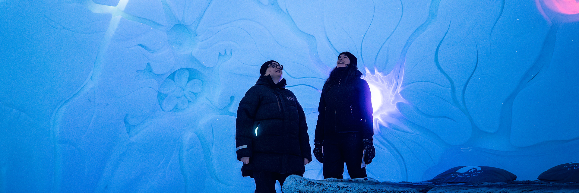 Visitors admiring carved ice wall art inside an ice hotel gallery, lit in blue and purple.