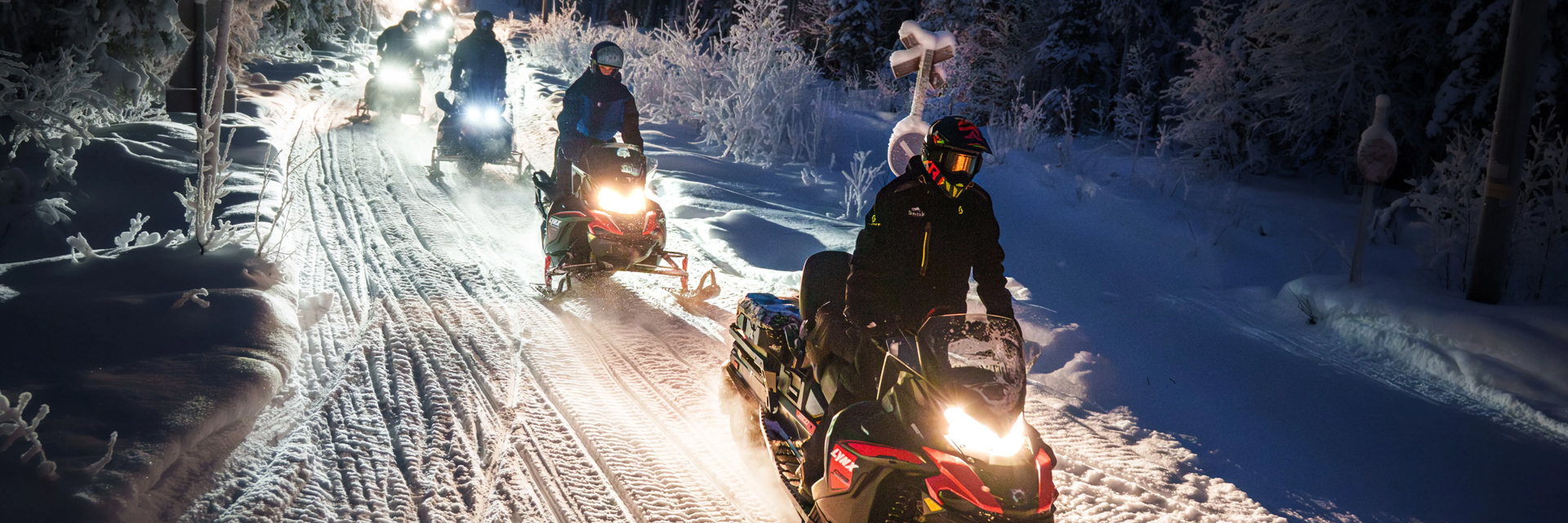 A convoy of snowmobiles moves in single file through a frost-covered forest trail at night in Lapland.