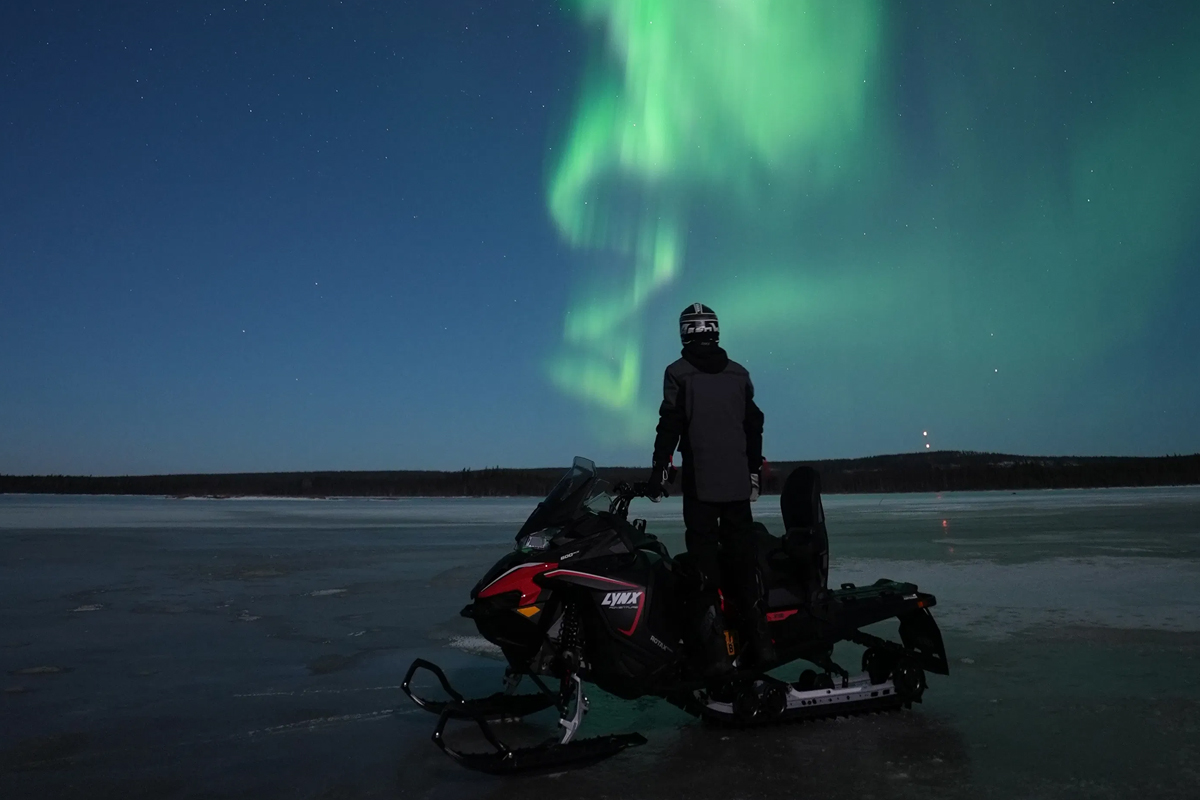 A rider stands beside a Lynx snowmobile on a frozen lake, watching the northern lights dance overhead in Lapland.
