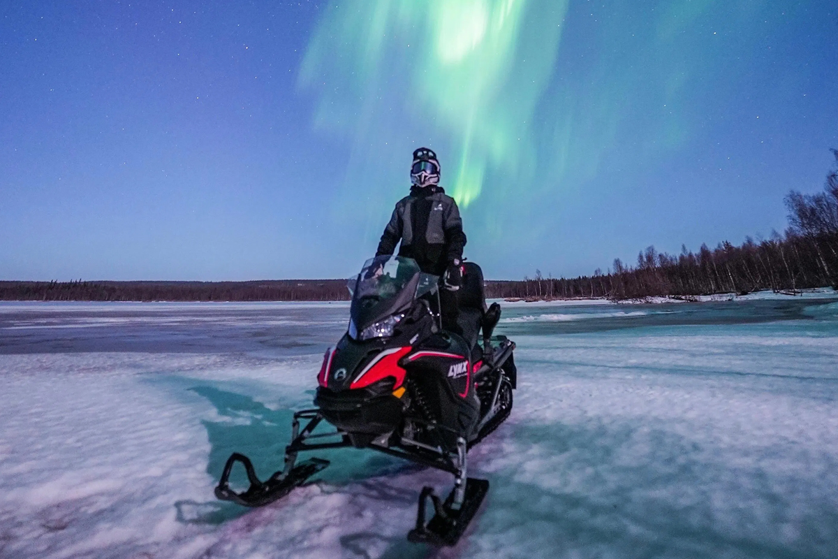 A rider on a Lynx snowmobile pauses on a frozen lake under vivid green northern lights in Lapland.