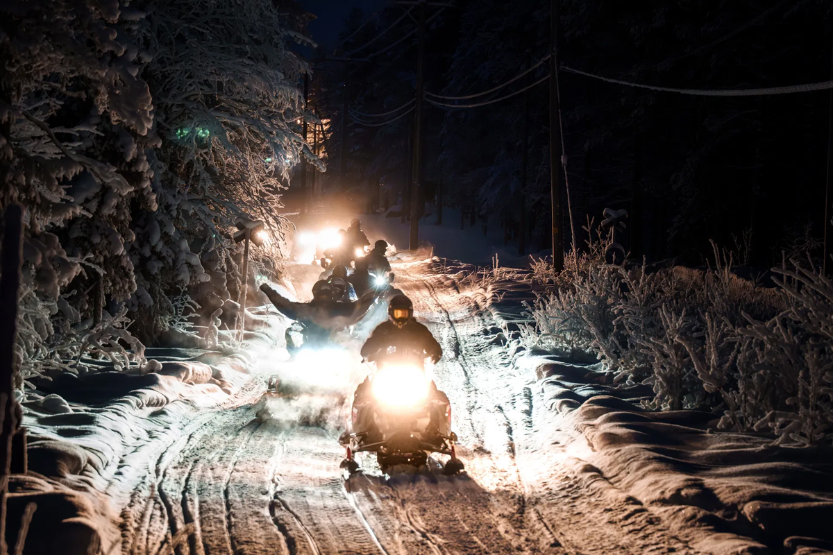 A group of snowmobiles rides through a dark forest trail at night, headlights cutting through the snow in Lapland.