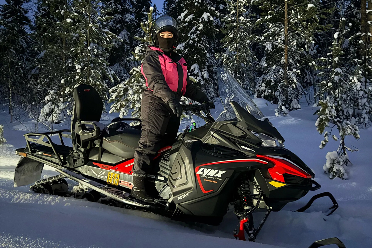 A rider in a pink jacket stands on a Lynx snowmobile on a snow-covered forest trail during a snowmobile safari in Lapland.