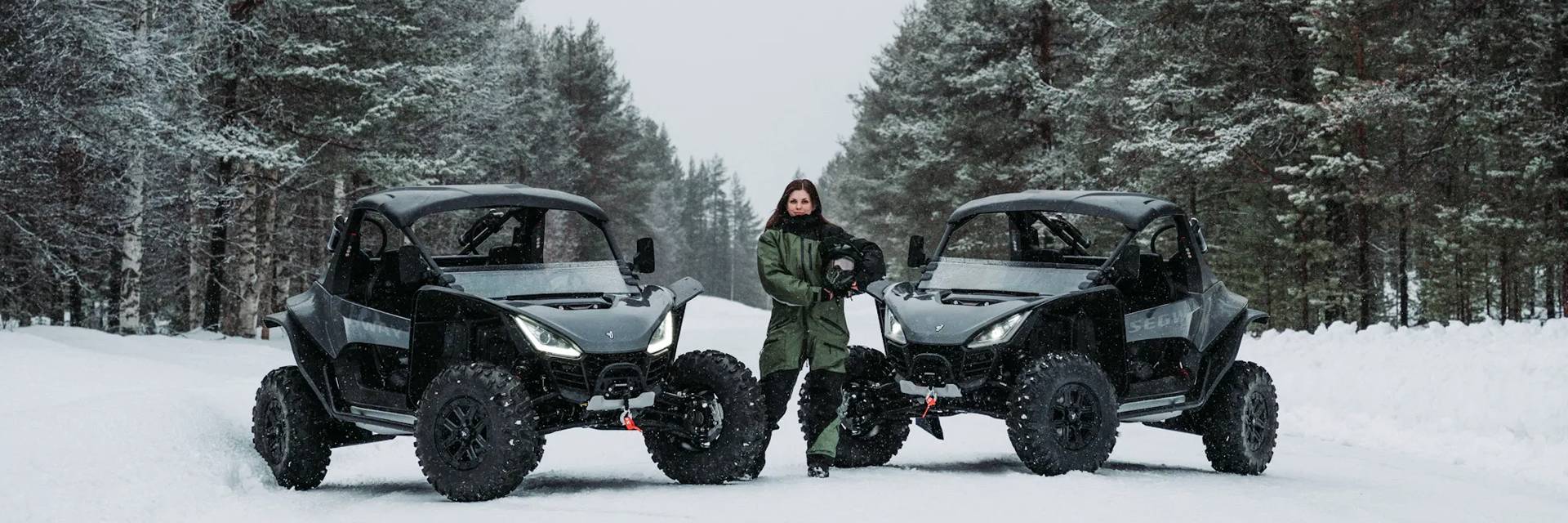 A woman in a green suit stands between two black ATV buggies on a snowy forest road in Lapland.
