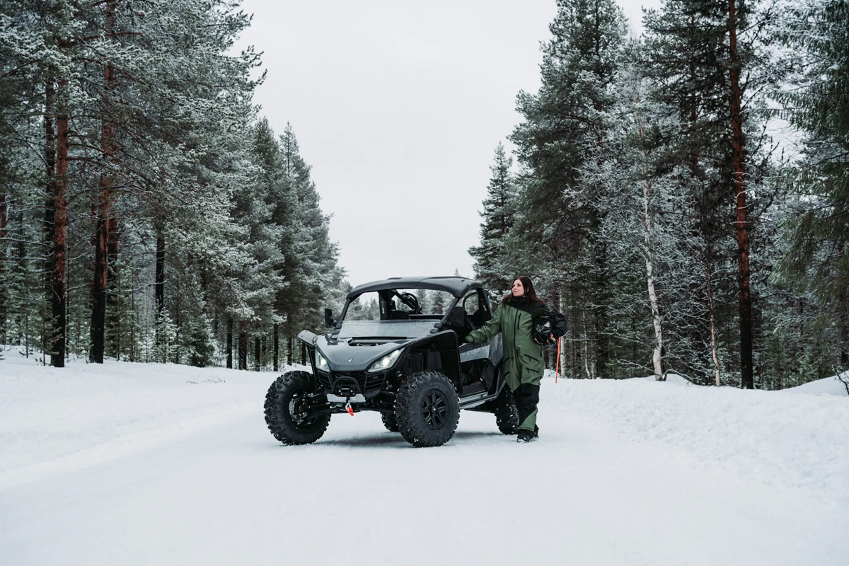 A woman in a green suit stands beside a black ATV buggy on a snowy forest trail in Lapland.