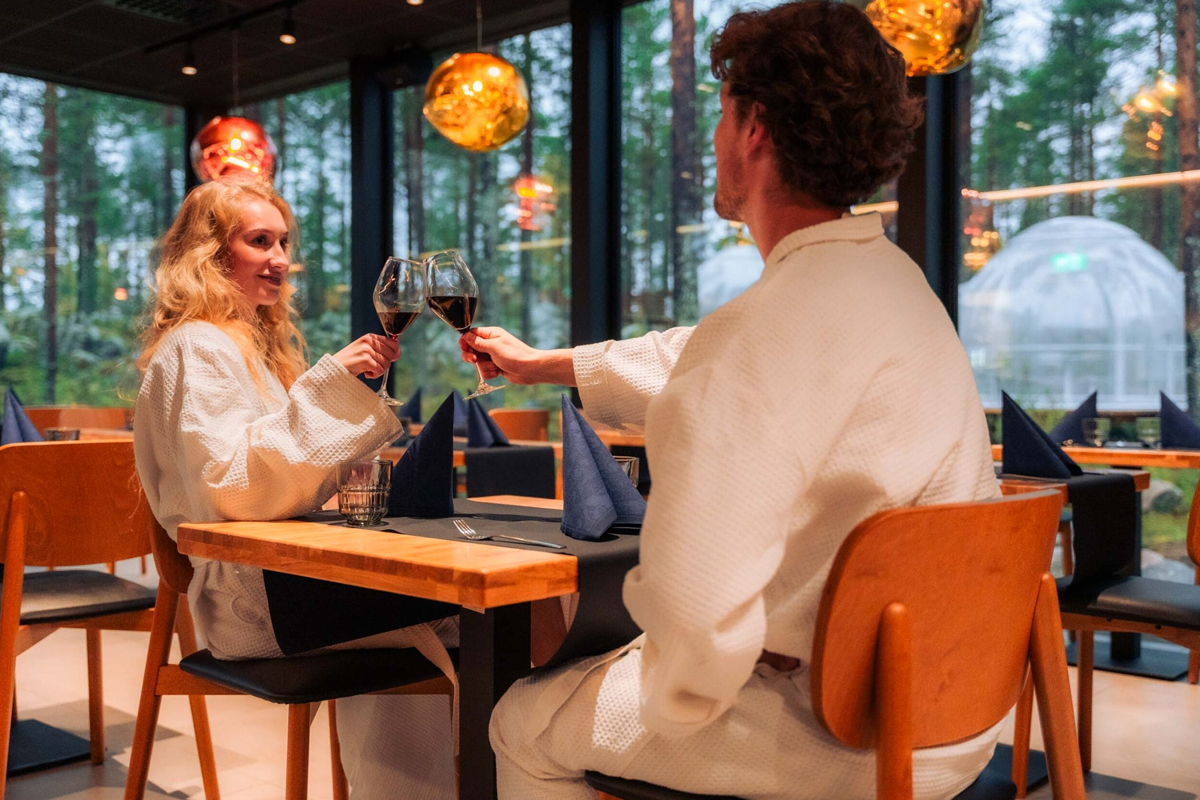 Couple in bathrobes enjoying dinner at Arctic Forest Spa, with glass igloos visible outside.