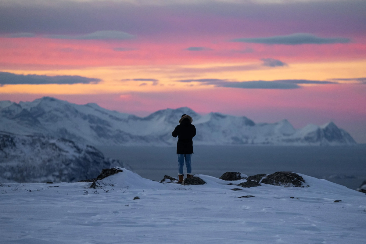 Traveler standing on a snowy ridge at sunset above an Arctic fjord. Photo: Northern Soul Adventures.