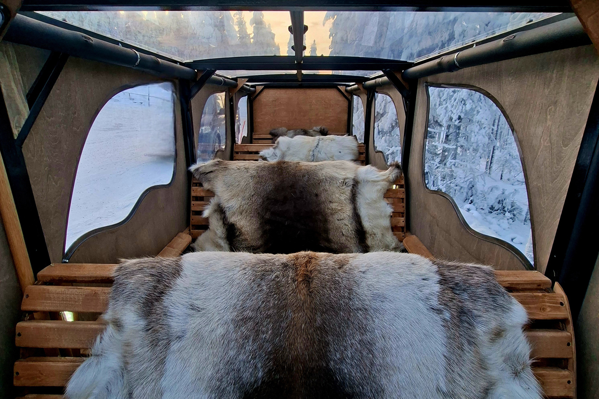 Interior of a heated sleigh with wooden bench seats and reindeer fur throws, windows overlooking a snowy forest