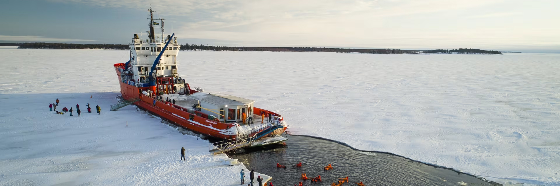 Icebreaker ship Polar Explorer cutting through frozen sea, with guests floating in survival suits nearby.